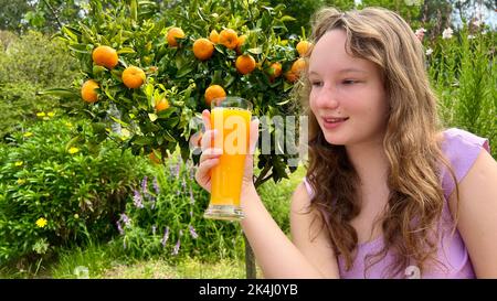 la ragazza beve succo d'arancia sullo sfondo di un albero di mandarino, può essere succo d'arancia mango mandarino beve avidamente e ama davvero il succo delizioso ovunque verdi ed estate. Foto Stock