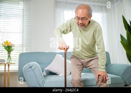 Anziano asiatico con tipo di occhiali in piedi dal divano con bastone di canna da camminata per camminare a casa, anziani affetti da dolore al ginocchio tenere Foto Stock