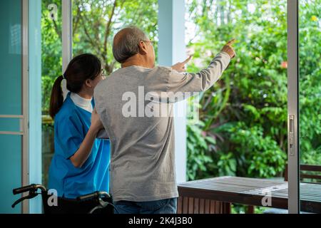 Giovane infermiera asiatica sta sostenendo l'uomo anziano seduto in sedia a rotelle a piedi guardando fuori finestra sul giardino e respirare aria fresca, pensione CE Foto Stock