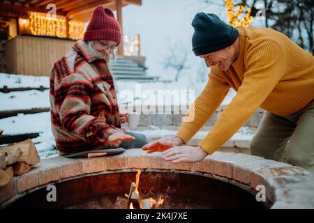 Coppia anziana seduta e riscaldamento insieme al caminetto all'aperto nel loro giardino d'inverno. Foto Stock