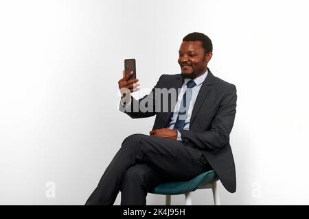 Sorridente uomo nero afro-americano in tuta da lavoro seduto su sedia fare videochiamata via smartphone, sfondo bianco parete. Ritratto a grandezza naturale di ha Foto Stock
