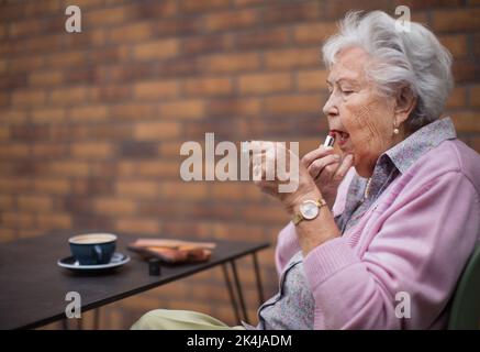 Donna anziana felice che mette il rossetto sulle sue labbra godendosi il caffè in città. Foto Stock