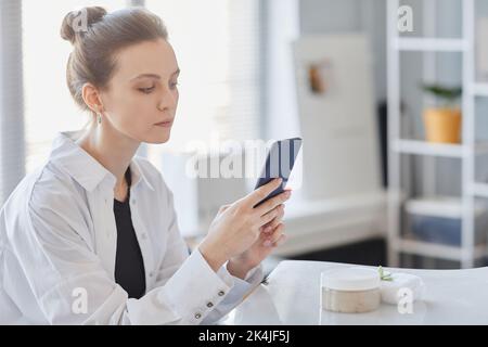 Vista laterale ritratto di giovane donna che prende immagini di prodotto di crema di bellezza fatta a mano con smartphone Foto Stock