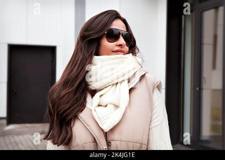 Primo piano Ritratto di bella donna che guarda via sulla strada. Modello elegante con lunghi capelli brunette che indossano occhiali da sole, sciarpa bianca lavorata a maglia e maglione Foto Stock