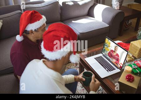 Papà biraciale e figlio adulto in cappelli di babbo natale laptop videochiamata di natale con donna felice. natale, festa e tecnologia di comunicazione. Foto Stock