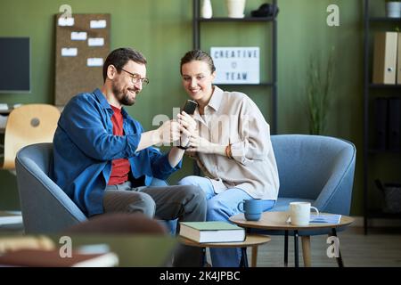 Felice giovane uomo d'affari con lo smartphone che mostra video online ai colleghi sulle nuove tendenze, cercando allo stesso tempo nuove idee per il progetto Foto Stock