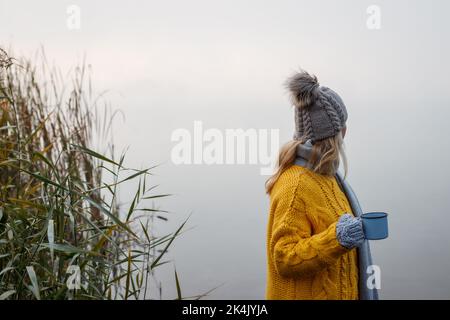 Donna che si rilassa con una bevanda calda accanto al lago in autunno mattina fredda. Campeggio all'aperto nella stagione autunnale. Donna bionda con cappello a maglia e sudore giallo Foto Stock