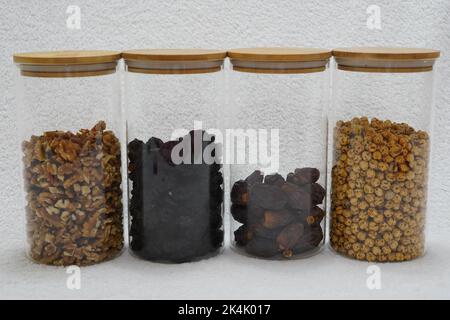 prugne secche, datteri, ceci, noci e anacardi in vasetti di vetro con coperchi di legno su fondo bianco in rustico cucina bianca Foto Stock