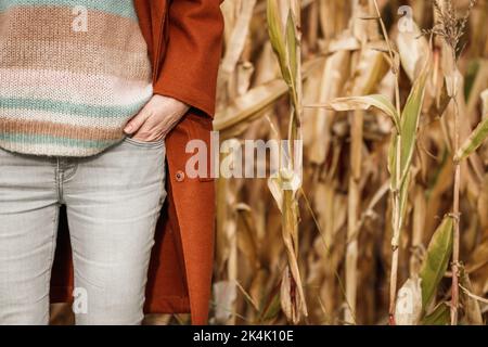 Donna con cappotto rosso, jeans skinny e maglione di lana in piedi in campo di mais all'aperto. Abbigliamento autunnale Foto Stock