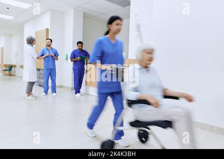 i medici parlano, l'infermiera sta spingendo la sedia a rotelle con pazienti anziani in movimento, il personale medico sta lavorando nella hall dell'ospedale. Istituzione sanitaria Foto Stock
