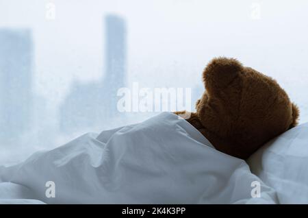 Orsacchiotto che dorme da solo sul letto con cuscino bianco e coperta che si affaccia sulla finestra con caduta di pioggia nel giorno solitario. Foto Stock