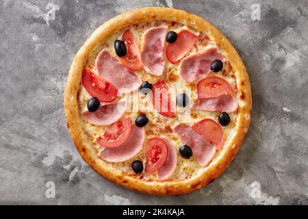 Pizza classica sottile con formaggio fuso, prosciutto, pomodori e olive nere su superficie di pietra grigia Foto Stock