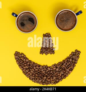 Un'emoticon sorridente, le labbra fatte di chicchi di caffè e gli occhi fatti di tazze di caffè. Foto Stock
