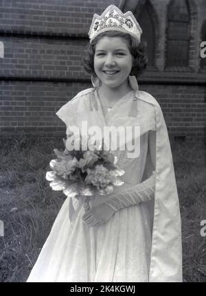 1959, storica, una ragazza adolescente, la locale May Queen, in piedi per la sua foto nel suo lungo abito, con fiori e corona sulla testa, Leeds, Inghilterra, Regno Unito. La sua foto è scattata nei giardini della chiesa prima del carnevale, dove sarà una figura di spicco nelle tradizionali celebrazioni del giorno di maggio, un'antica festa europea che segna l'inizio dell'estate. Foto Stock