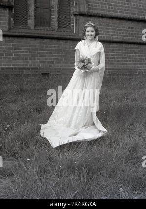 1959, storica, una ragazza adolescente, la locale May Queen, in piedi per la sua foto nel suo lungo abito, con fiori e corona sulla testa, Leeds, Inghilterra, Regno Unito. La sua foto è scattata nei giardini della chiesa prima del carnevale, dove sarà una figura di spicco nelle tradizionali celebrazioni del giorno di maggio, un'antica festa europea che segna l'inizio dell'estate. Foto Stock