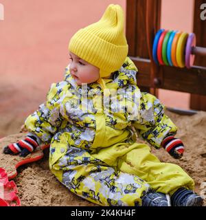 Il bambino felice del bambino del bambino sta giocando nel sandbox. Un bambino in abiti caldi gioca con la sabbia nel parco giochi. Bambino di un anno e due mesi in Foto Stock