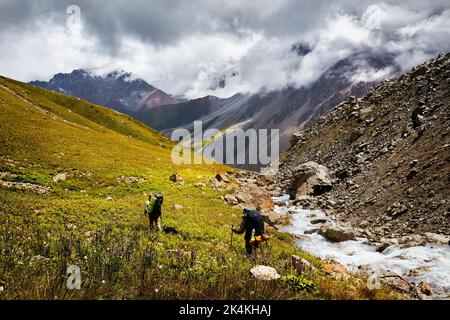 Due escursionisti uomini turisti con zaino e cane a piedi sulla collina vicino al fiume contro la nebbia e cielo scuro sovrastato nella valle di montagna. All'aperto e trekking Foto Stock