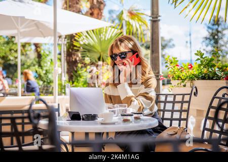 Foto di una donna felice di mezza età che chiama mentre si siede al bar all'aperto. Uomo d'affari sicuro che indossa abiti casual e che usa un computer portatile durante la marcia Foto Stock