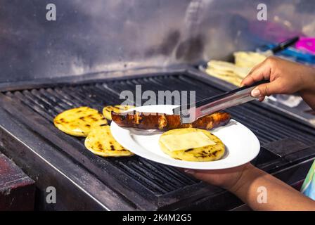 Mani di donna che servono arepas con formaggio e chorizo su un piatto accanto alla griglia. Cibo popolare colombiano Foto Stock