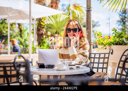 Foto di una donna felice di mezza età che chiama mentre si siede al bar all'aperto. Uomo d'affari sicuro che indossa abiti casual e che usa un computer portatile durante la marcia Foto Stock