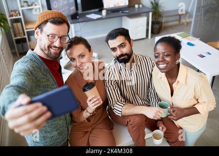 Gruppo di quattro giovani colleghi multiculturali con tazze di caffè e tè che fanno selfie in pausa, mentre tre di loro seduti sulla scrivania Foto Stock