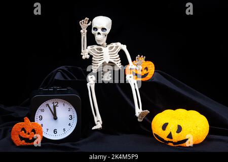 Halloween. Uno scheletro che striscio fuori dalla tomba e agita la mano, saluto, zucche con le grimaces, un orologio su sfondo nero. La vigilia di Foto Stock