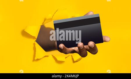 Mano maschio nera che tiene la carta di credito della banca nel foro strappato di fondo giallo, panorama, primo piano Foto Stock