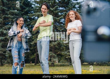 Gruppo di tre influencer che girano video di danza per il social network sul cellulare, divertendosi all'aperto. I blogger millennial trasmettono contenuti in streaming per loro Foto Stock