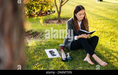 Giovane donna seduta in un parco su un prato e prendere appunti in un notebook. La ragazza lavora, studia, impara, tiene un diario. Filtro Instagram. Stile di vita Foto Stock