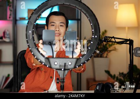 Focus selettivo del giovane influencer maschile asiatico che si prepara a fare contenuti per blog impostazione di due smartphone a stand lampada ad anello Foto Stock