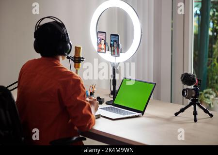 Fuoco selectiv di moderno giovane adulto asiatico maschio influencer seduto alla scrivania di fronte alla lampada ad anello con due smartphone in streaming dal vivo Foto Stock