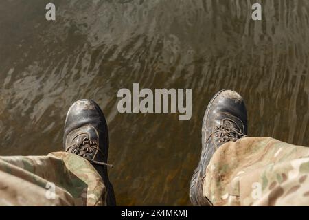 Sezione bassa di persona sopra acqua. Le gambe da uomo in pantaloni camouflage e vecchi stivali dangolano sopra l'acqua. Foto Stock