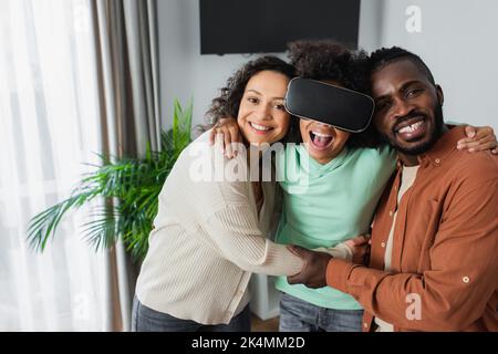 felici genitori afroamericani che abbraccia entusiasti giochi di bambina in visore vr, immagine stock Foto Stock