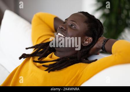 Primo piano Ritratto di Dreamy Black Man che si appoggia sul divano e sorridente Foto Stock