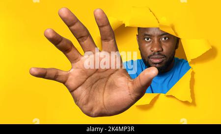 Serio uomo afroamericano che raggiunge attraverso il foro in carta gialla strappata, cercando di afferrare qualcosa Foto Stock