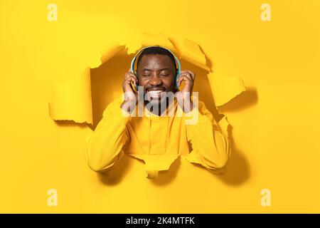 Uomo afroamericano soddisfatto che sbirciava fuori buco su sfondo giallo e ascoltando musica in cuffie Foto Stock