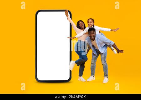 Felice eccitato african american coppia millennial con bambina divertirsi vicino grande smartphone con schermo vuoto Foto Stock