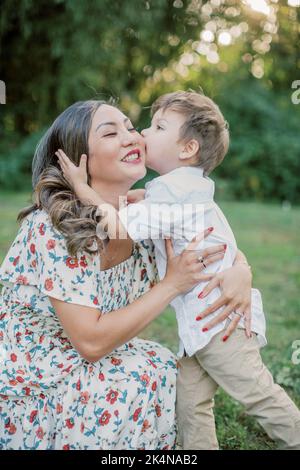 Il ragazzo di 2-4 anni bacia la guancia di sua madre asiatica fuori. Foto Stock