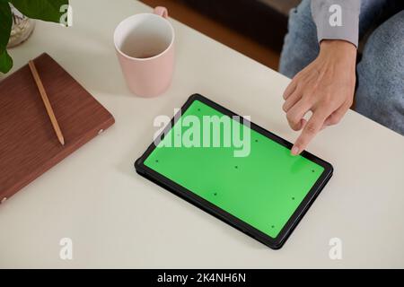 Vista dall'alto della mano di una giovane donna che indica lo schermo verde vuoto del tablet mentre si siede al tavolo e presenta una nuova applicazione Foto Stock