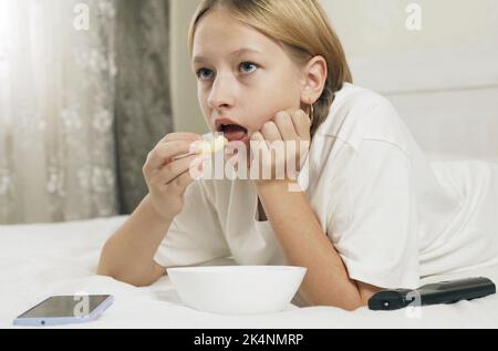 Una ragazza adolescente si trova su un letto, mangia bastoni di mais e guarda in uno smartphone e TV. Primo piano. Foto Stock