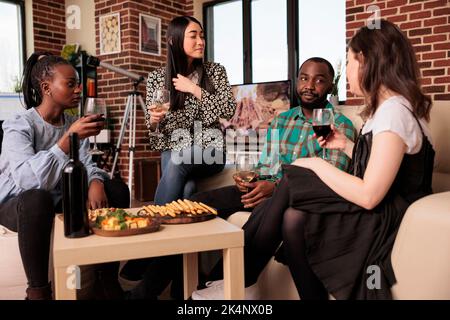 Culturalmente multi donne diverse bere vino, mangiare grissini riuniti in afro americano amico maschio home.varie nazionalità partner conversazione, parlare, chiacchierare a snack partito. Foto Stock