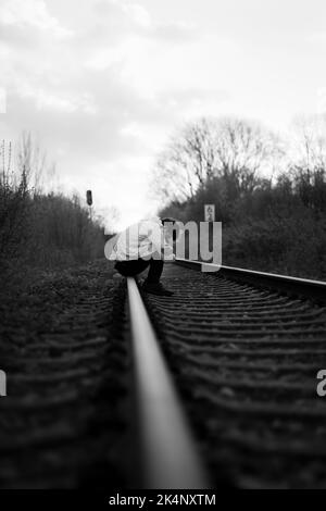 Un colpo verticale in scala di grigi di un uomo triste seduto sulle piste del treno - concetto di depressione Foto Stock