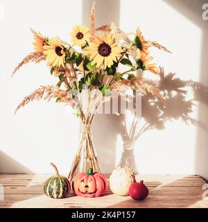 Girasoli in vaso di vetro con piccole zucche decorative contro la parete bianca alla luce del sole. Concetto di ringraziamento. Ancora vita. Primo piano. Foto Stock