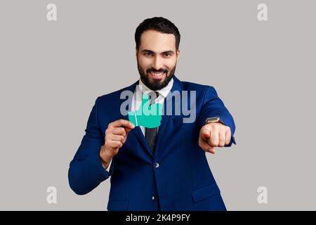 Ritratto di bell'uomo d'affari bearded entusiasta che punta alla macchina fotografica con il sorriso e che mostra il pollice della carta in su, portando il vestito ufficiale di stile. Studio in interni isolato su sfondo grigio. Foto Stock