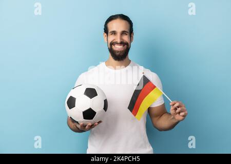 Ritratto di uomo sorridente con barba che indossa una T-shirt bianca a sostegno della squadra di calcio tedesca in campionato, rallegrarsi e salutare, patriottismo. Studio in interni isolato su sfondo blu. Foto Stock