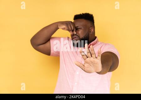 Ritratto dell'uomo pizzicando il naso, smettere di respirare cattivo odore, disgustato da odore di farting, esprimendo repulsione, grossolana, mostrando stop gesto con palmo. Studio al coperto isolato su sfondo giallo. Foto Stock