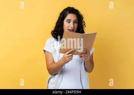 Ritratto di stupito impressionato donna sorpresa con i capelli ondulati scuri in piedi con il libro nelle mani, leggendo romanzo stupefacente, guardando le pagine con gli occhi grandi. Studio al coperto isolato su sfondo giallo. Foto Stock