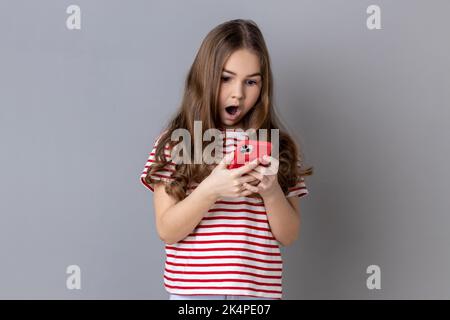 Ritratto di una bambina stupefacente che indossa una T-shirt a righe utilizzando uno smartphone, guarda lo schermo del dispositivo, scrive post per il blog del suo bambino. Studio in interni isolato su sfondo grigio. Foto Stock