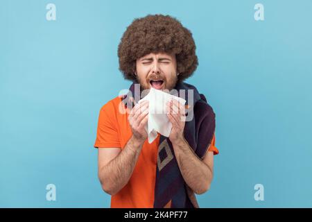 Uomo con acconciatura afro avvolto in tessuto sciarpa starnuti, tosse, sensazione di malessere, malessere o influenza, allergia o sintomi influenzali stagionali. Studio in interni isolato su sfondo blu. Foto Stock