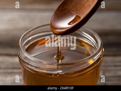 Miele e cucchiaio di legno in contenitore di vetro su vecchio sfondo di legno. Miele che sgocciola dal cucchiaio. Foto Stock